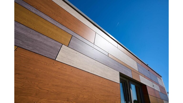 Bunte Fassadenverkleidung mit Holzoptik-Paneelen in verschiedenen Farbtönen, aufgenommen bei klarem, sonnigem Himmel.